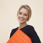 A cheerful woman smiles while holding an orange folder against a neutral background.