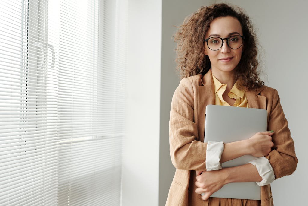 Curly-haired businesswoman wearing glasses holds a laptop in a bright office setting.